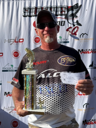 Man holding a trophy and check in front of sponsor backdrop, wearing sunglasses and a cap