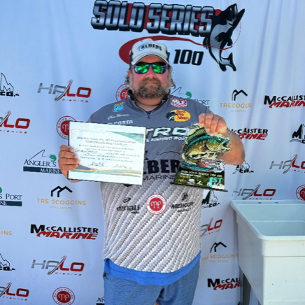Man standing on a sponsor backdrop, wearing a white fishing jersey and cap, holding a trophy plaque.