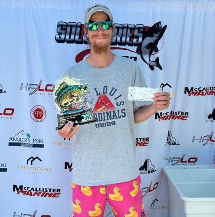 Man holding a check on a tournament stage with sponsor backdrop, wearing a gray shirt, cap, and sunglasses