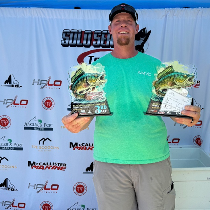 Angler holding a trophy and check at a fishing tournament podium, wearing a sponsor jersey and cap.