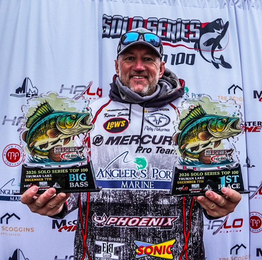 Man holding two trophy fish at a bass fishing awards backdrop, wearing a cap and smile.