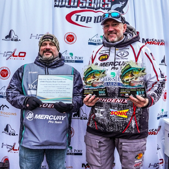 Two anglers in winter gear hold plaques and fish trophies on a Top 100 stage podium