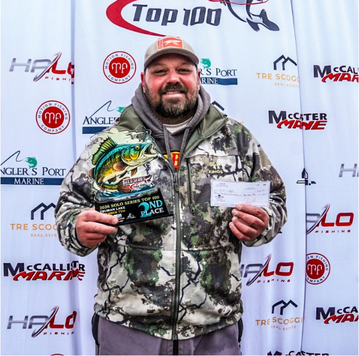 Man in camo jacket holding fish plaque and certificate in front of Top 100 sponsor backdrop