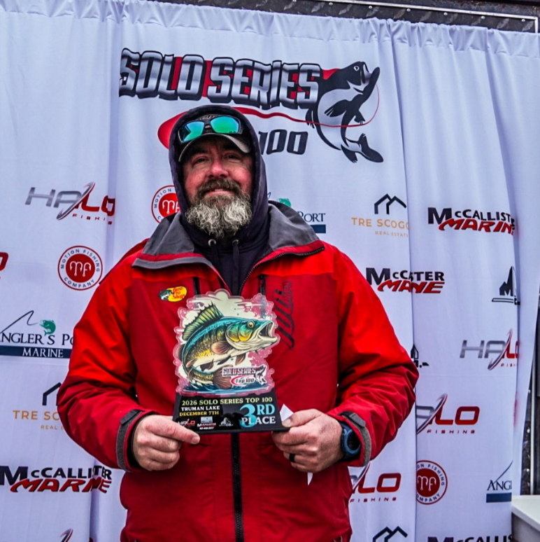 Man in red jacket holding an award plaque at a fishing event backdrop