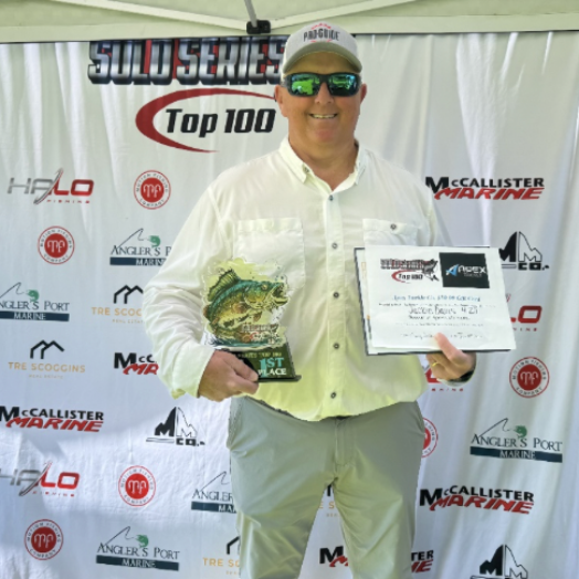 Man standing on a sponsor backdrop, wearing a white fishing jersey and cap, holding a trophy plaque.