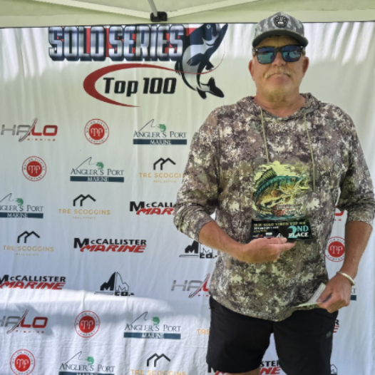 Man holding a check on a tournament stage with sponsor backdrop, wearing a gray shirt, cap, and sunglasses