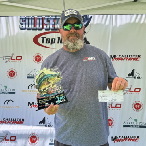 Angler holding a trophy and check at a fishing tournament podium, wearing a sponsor jersey and cap.
