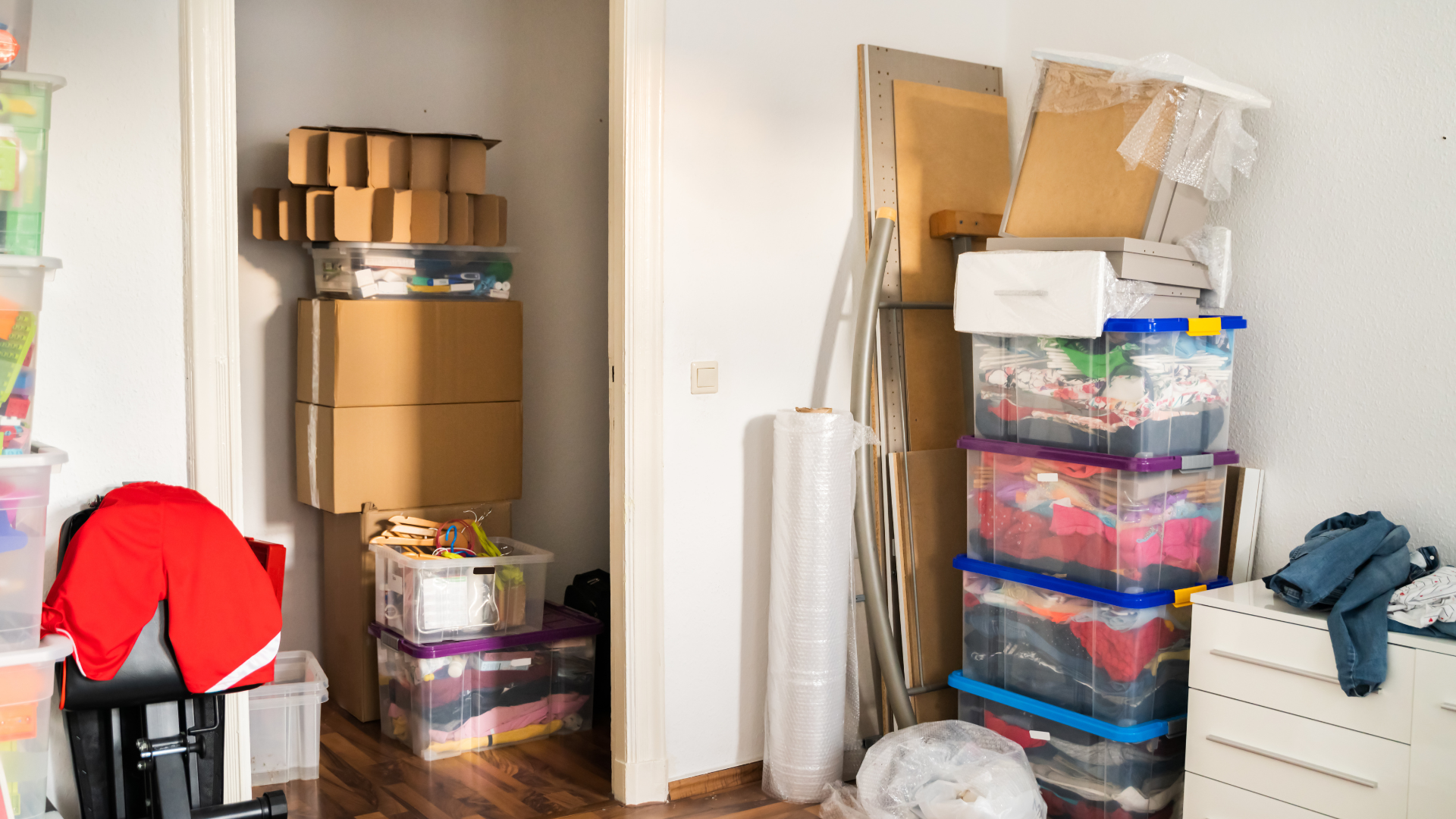 Cluttered storage room with stacked boxes, plastic bins, rolled materials, and a red jacket on a chair.