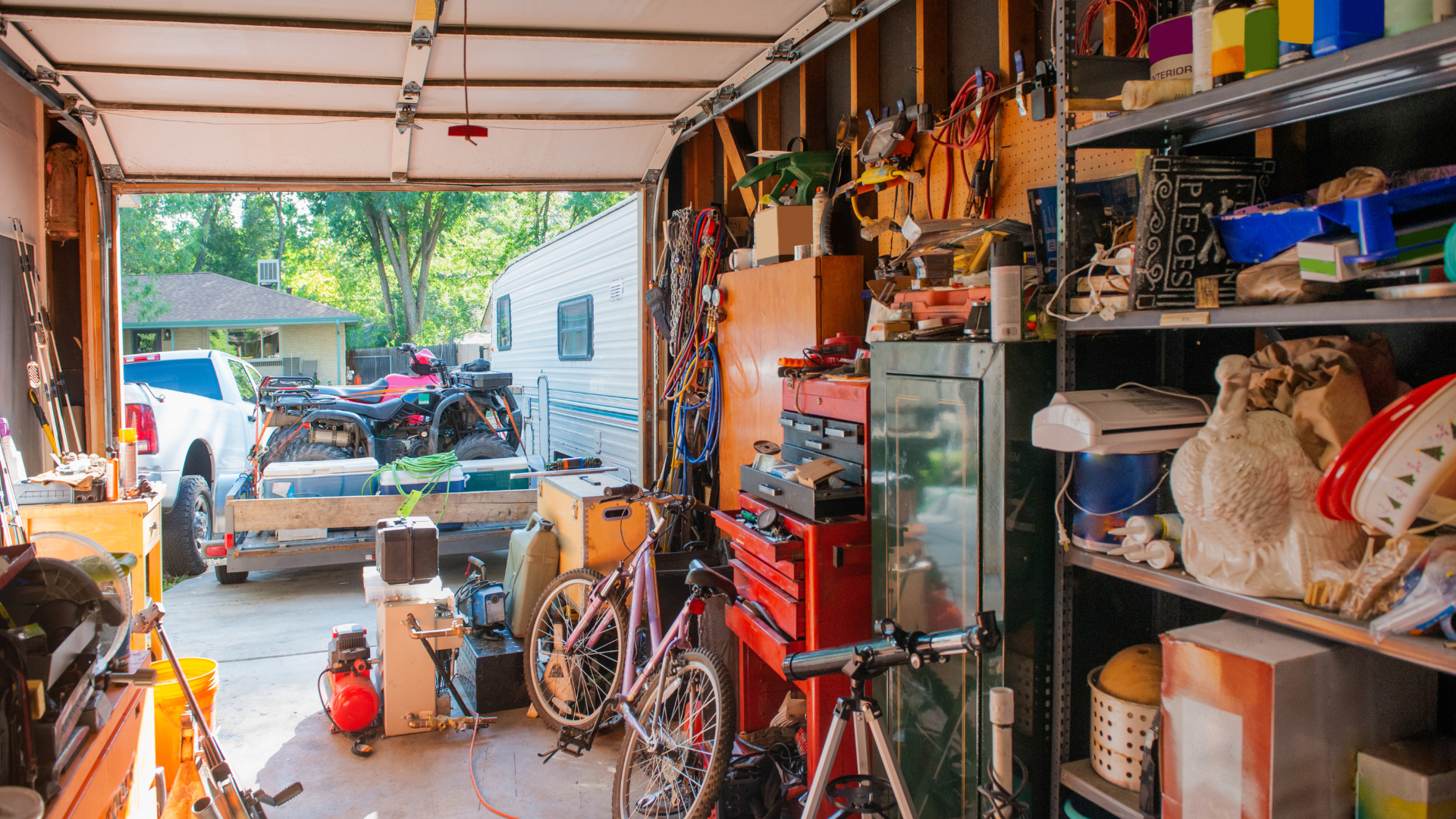 Garage cluttered with tools, shelves, and a workbench, looking out to a driveway with vehicles outside