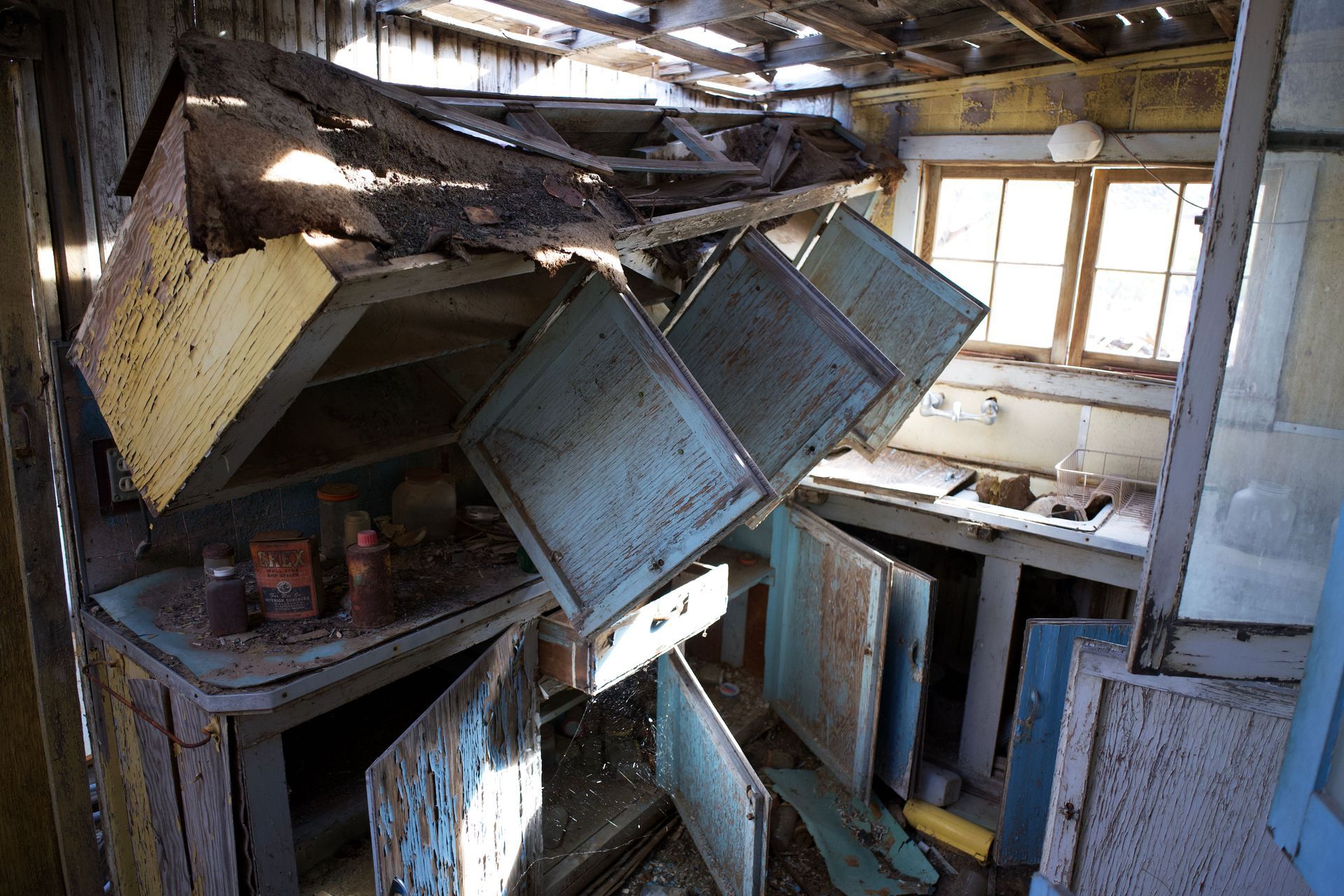 Collapsed, cluttered room with broken cabinets and debris under a damaged ceiling near windows.
