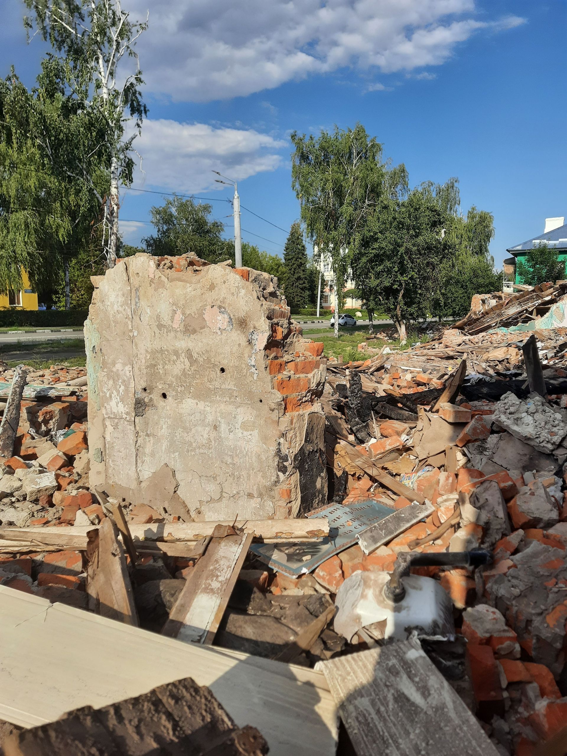 Collapsed brick ruins with a standing concrete pillar, scattered debris, and trees under a blue sky.