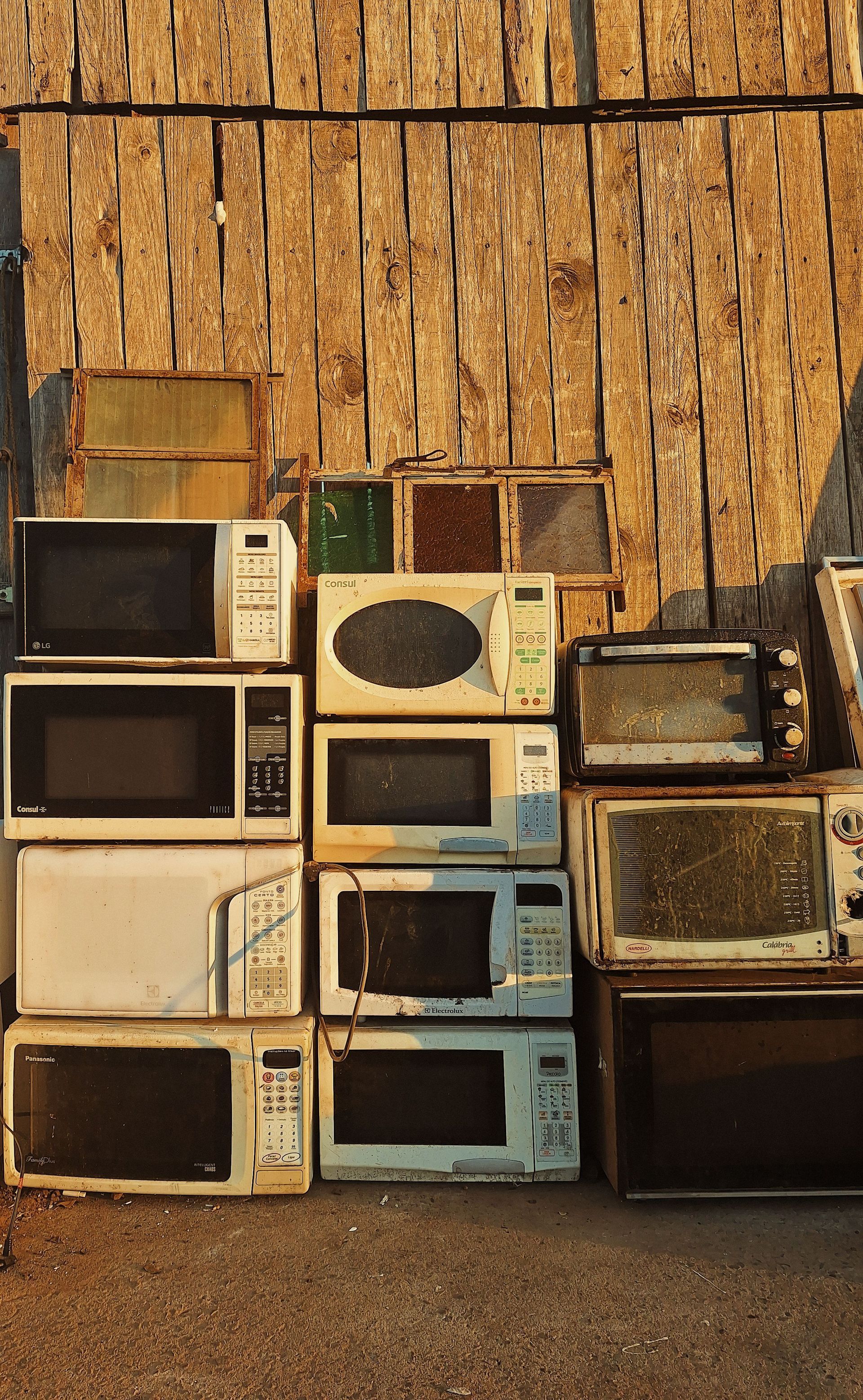 Stacked old microwaves and CRT TVs against a weathered wooden wall outdoors.