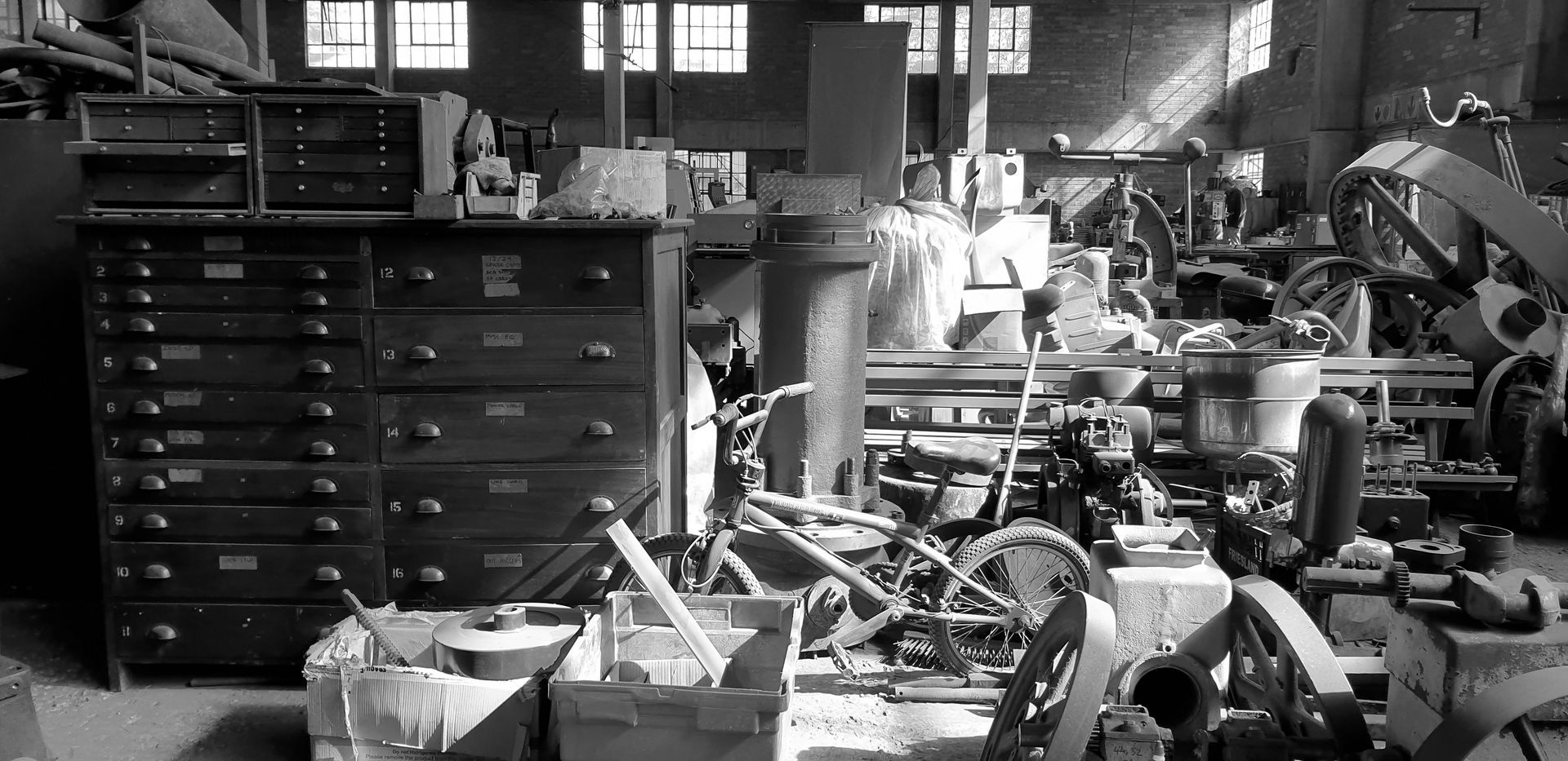 Black-and-white cluttered workshop with tool cabinets, machinery, and bicycles in a crowded garage.