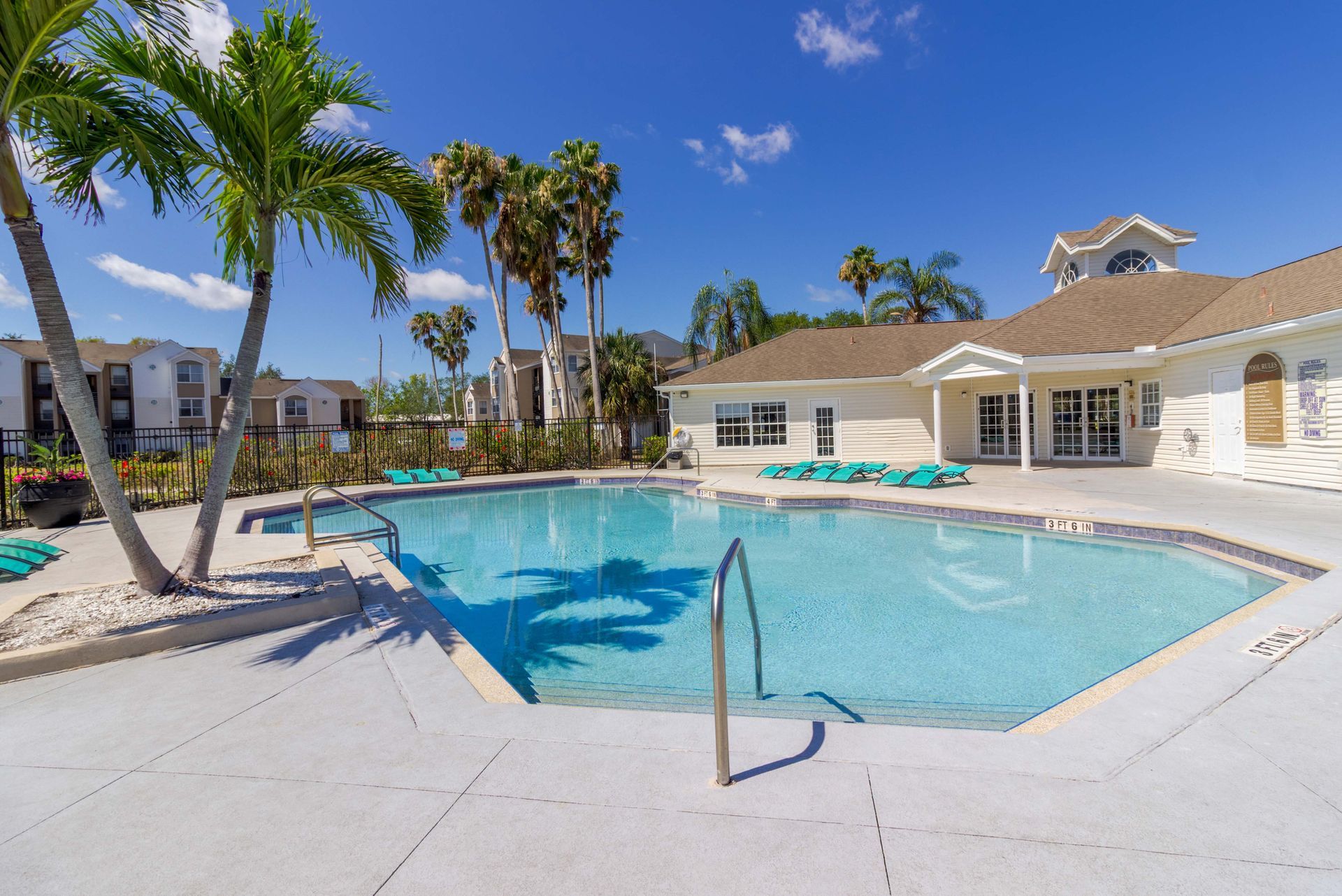 Resort-style pool with palm trees, lounge chairs, and a white clubhouse under a blue sky