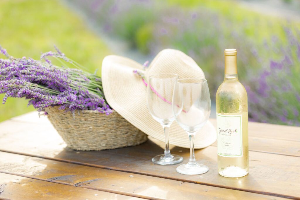 A bottle of wine , two wine glasses , a hat and a basket of lavender are on a wooden table.