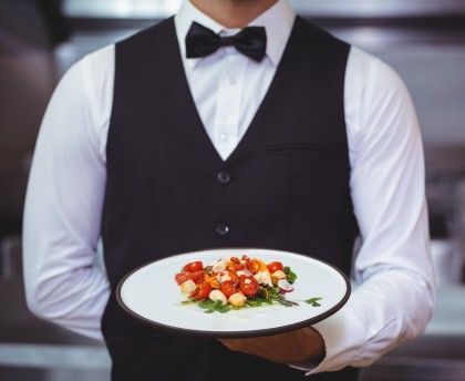 Camarero con chaleco negro sosteniendo un plato blanco con una colorida ensalada gourmet.
