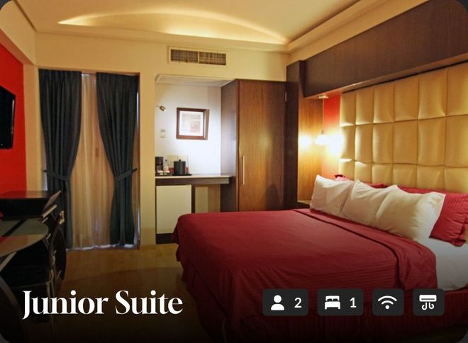 Suite junior de hotel con cama roja, cabecero acolchado, cortinas oscuras e iluminación ambiental cálida.
