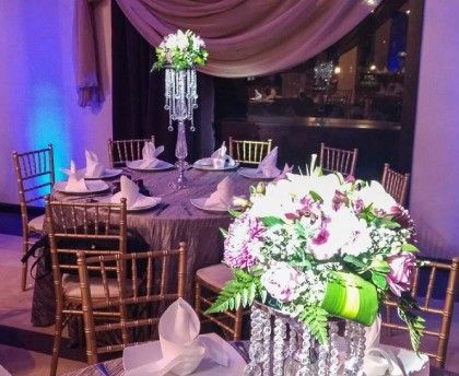 Elegante mesa de banquete con centros de mesa florales blancos y morados en un salón de eventos con iluminación tenue.