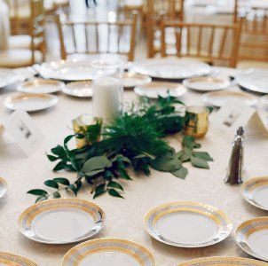 Juego de mesa formal con platos blancos, bajoplatos con borde dorado, centro de mesa con follaje y velas.