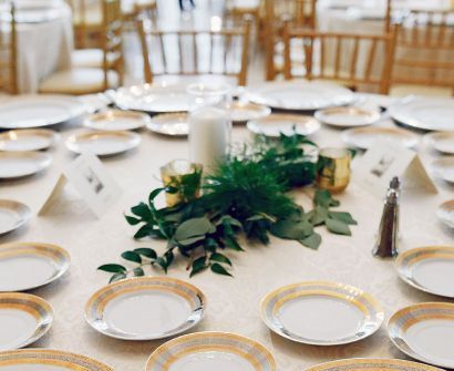 Elegante mesa de banquete con manteles blancos, platos con borde dorado, velas y centro de mesa con hojas verdes.