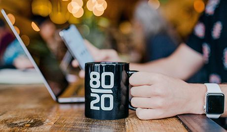Una mano sostiene una taza negra con la inscripción "80/20" junto a una computadora portátil en una mesa de café.