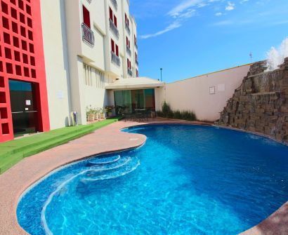 Piscina azul curvada junto a un edificio moderno con detalles rojos y un muro de piedra bajo un cielo despejado.