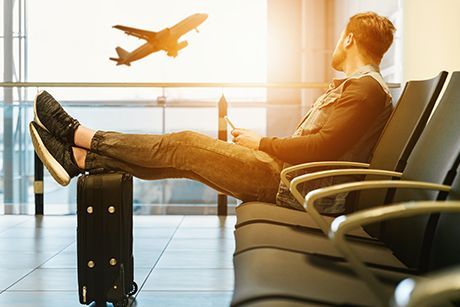 Un viajero recostado en un asiento del aeropuerto, mirando su teléfono mientras un avión despega al otro lado de la ventana.