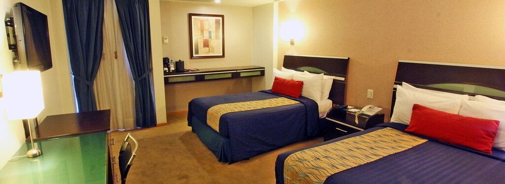 Habitación de hotel con dos camas, ropa de cama azul y roja, cortinas, lámpara y televisor sobre una cómoda.