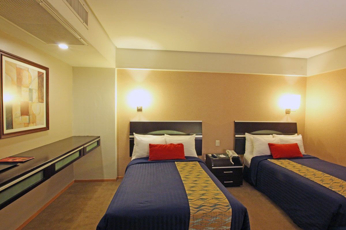 Habitación de hotel con una colcha roja, dos sillones, lámparas cálidas y suelo de baldosas.
