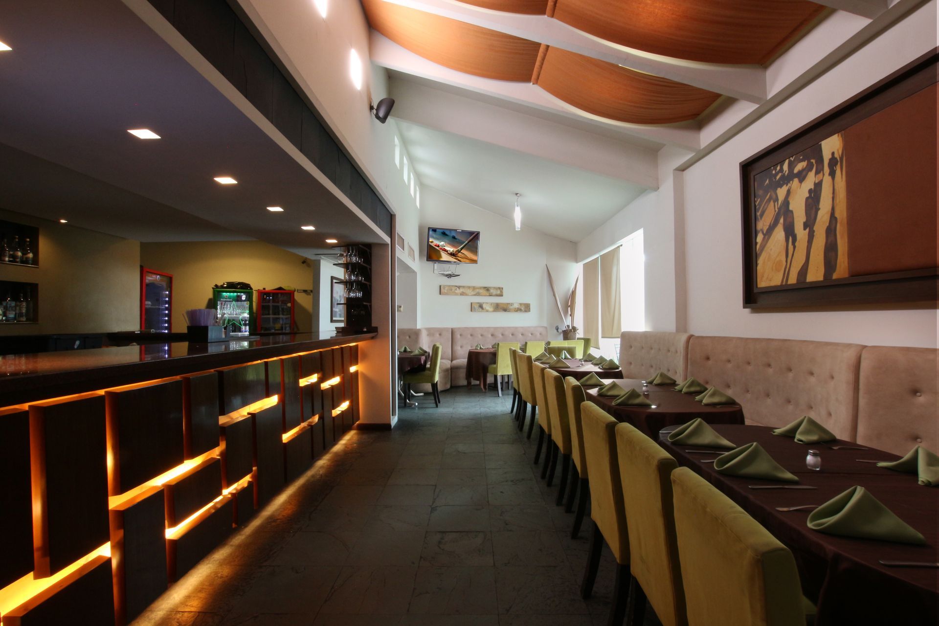 Interior de restaurante moderno y vacío con iluminación cálida, asientos en la barra y una larga mesa de comedor.