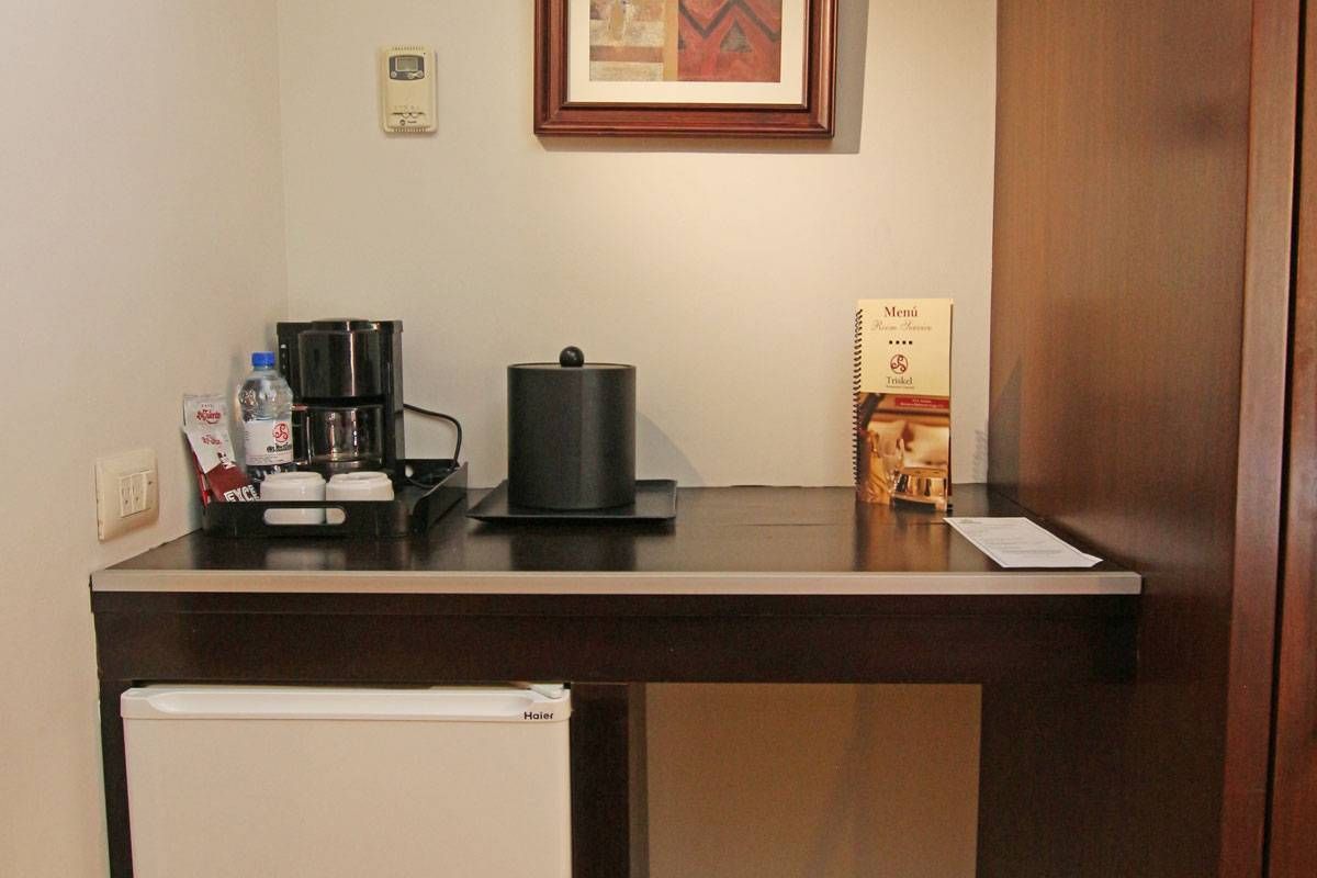 Escritorio pequeño en una habitación de hotel con cafetera, hervidor de agua, mininevera y cuadros enmarcados en la pared.
