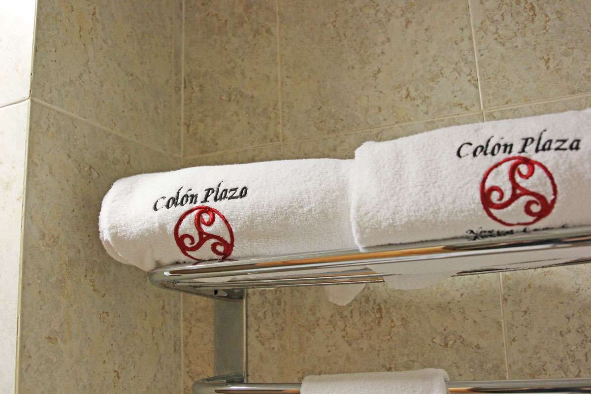 Toalla blanca bordada con el logotipo rojo de “Colón Plaza” en un soporte cromado sobre una pared de azulejos beige.