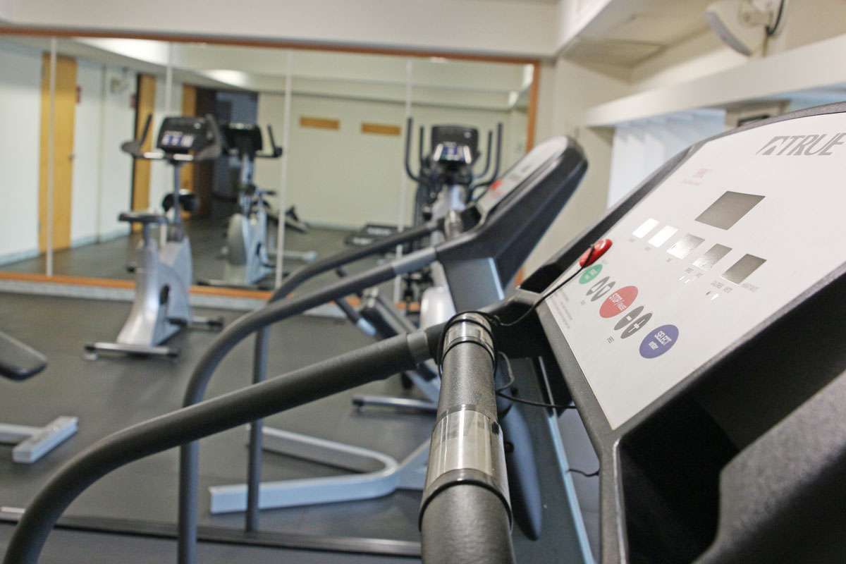 Máquinas de cardio de gimnasio con consola de cinta de correr en primer plano y bicicletas estáticas al fondo.