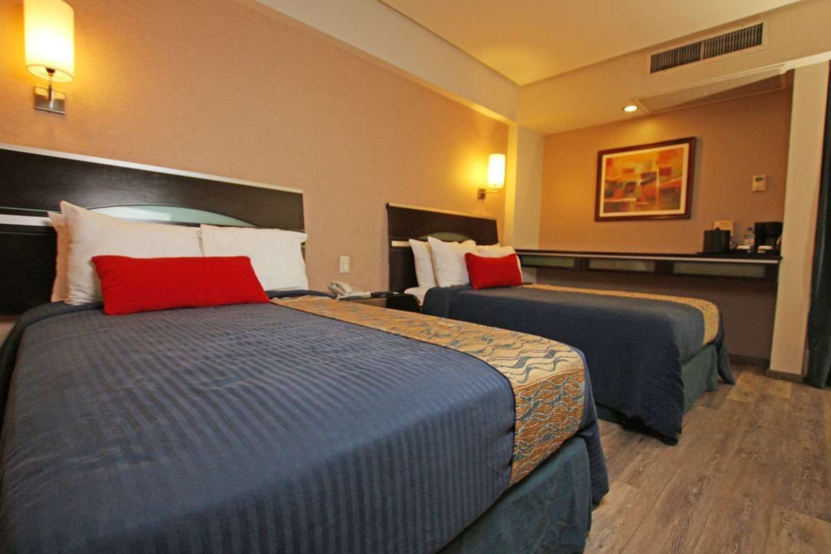 Habitación de hotel con dos camas dobles, ropa de cama azul, almohadas rojas y luces de pared cálidas.