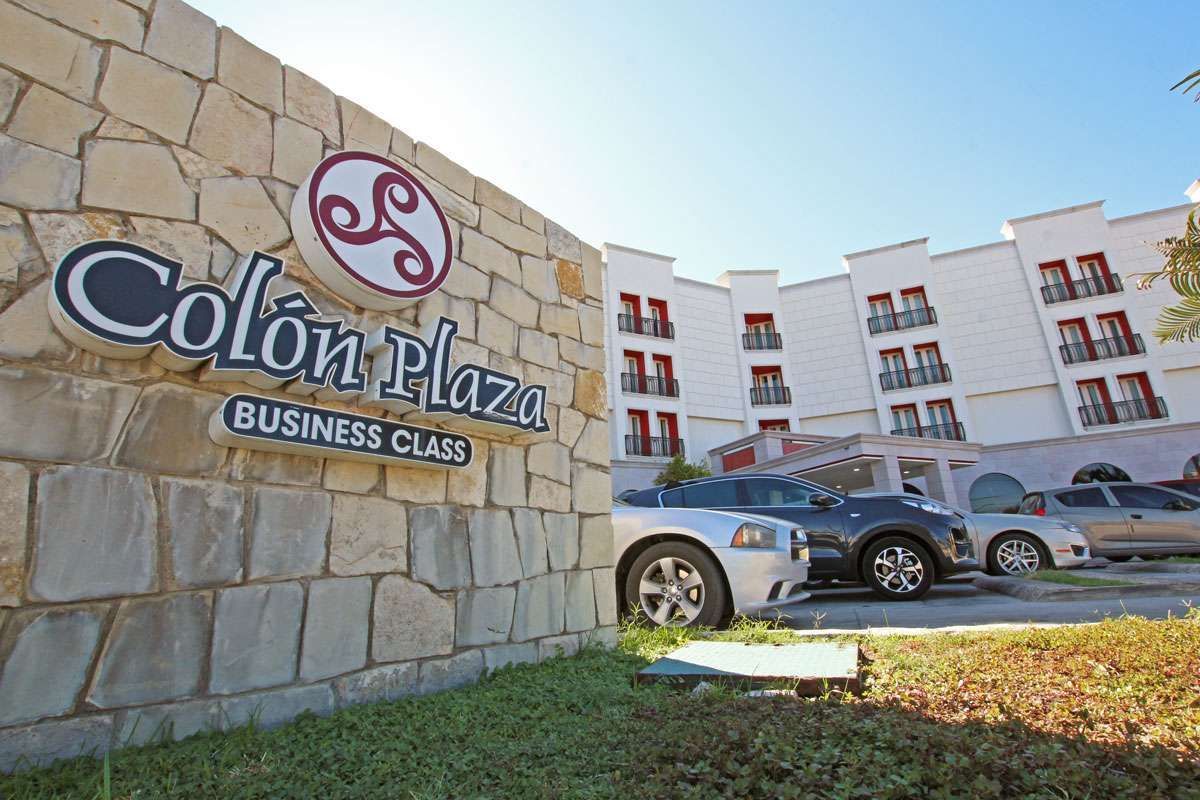 Letrero del club de negocios Colón Plaza junto a un muro de piedra y autos estacionados frente a un edificio blanco.