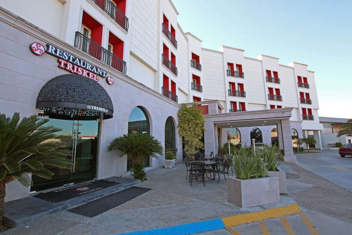 Entrada del hotel con fachada blanca, detalles en rojo, palmeras y asientos al aire libre bajo un toldo.