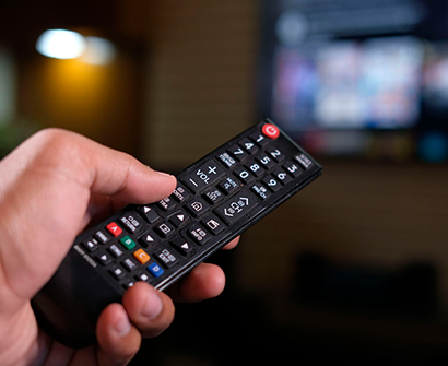 Una mano sostiene un control remoto negro frente a una pantalla de televisión borrosa.