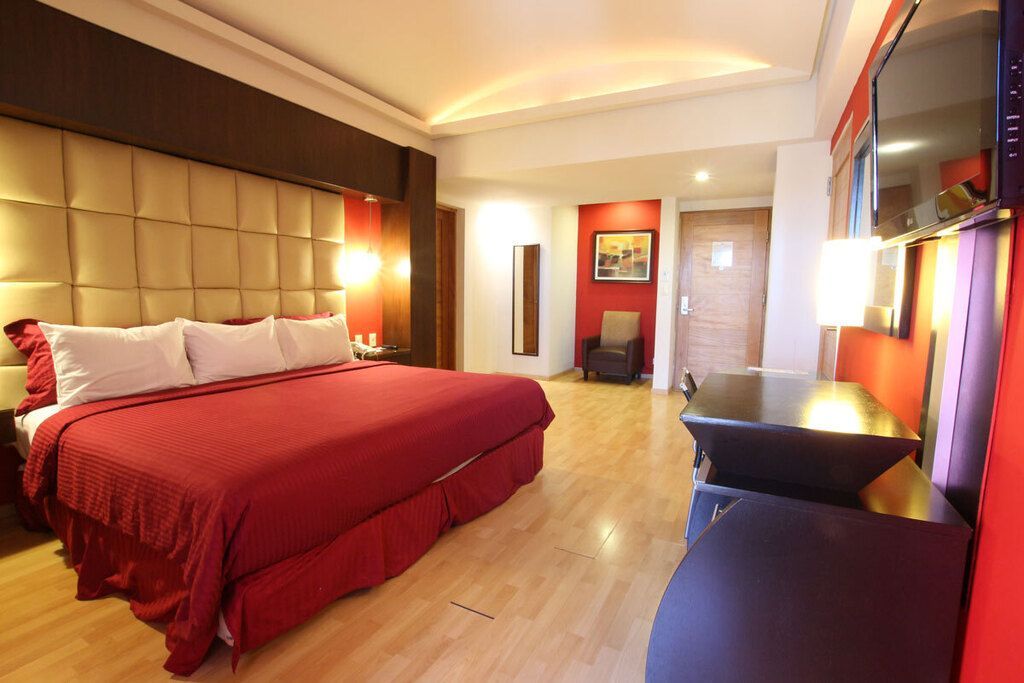 Habitación de hotel con una colcha roja, dos sillones, lámparas cálidas y suelo de baldosas.