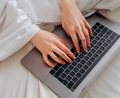 Manos tecleando en el teclado de un portátil sobre una cama, con una camisa blanca visible.