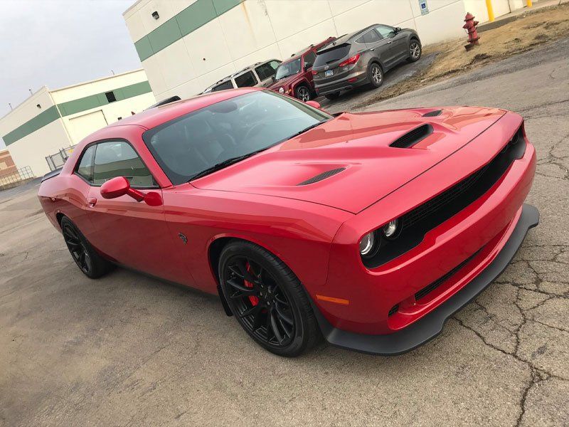 Red Dodge Challenger parked in front of a building. Black wheels, hood scoop, and spoiler. Gray asphalt.