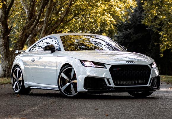 White Audi TT RS coupe parked on a road, trees in the background.