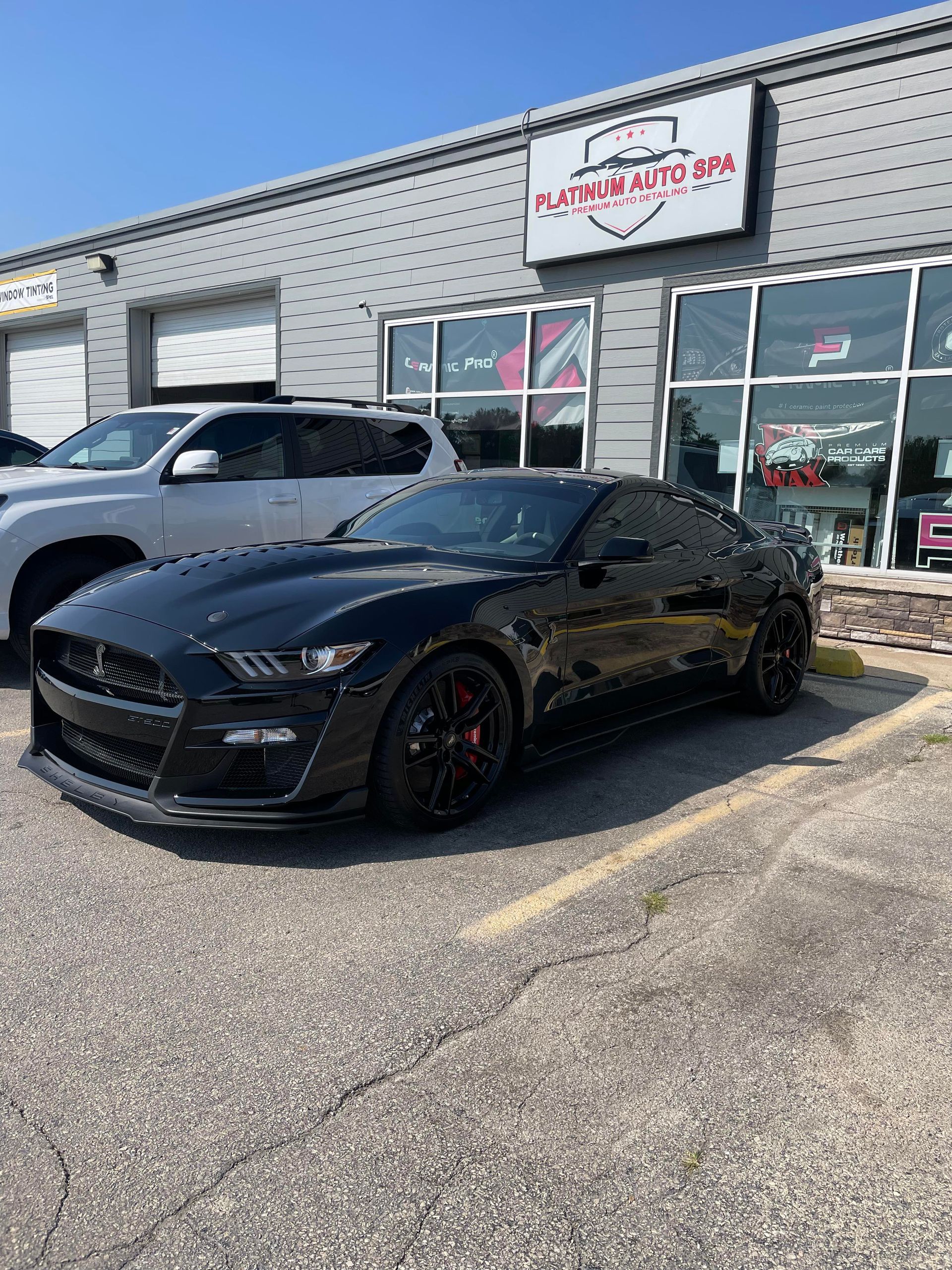 Black sports car parked outside an auto shop with white SUV in background on a sunny day.