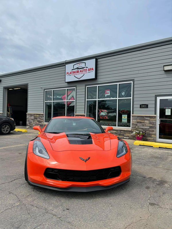 Orange Corvette in front of Platinum Auto Spa with a gray building and cloudy sky.