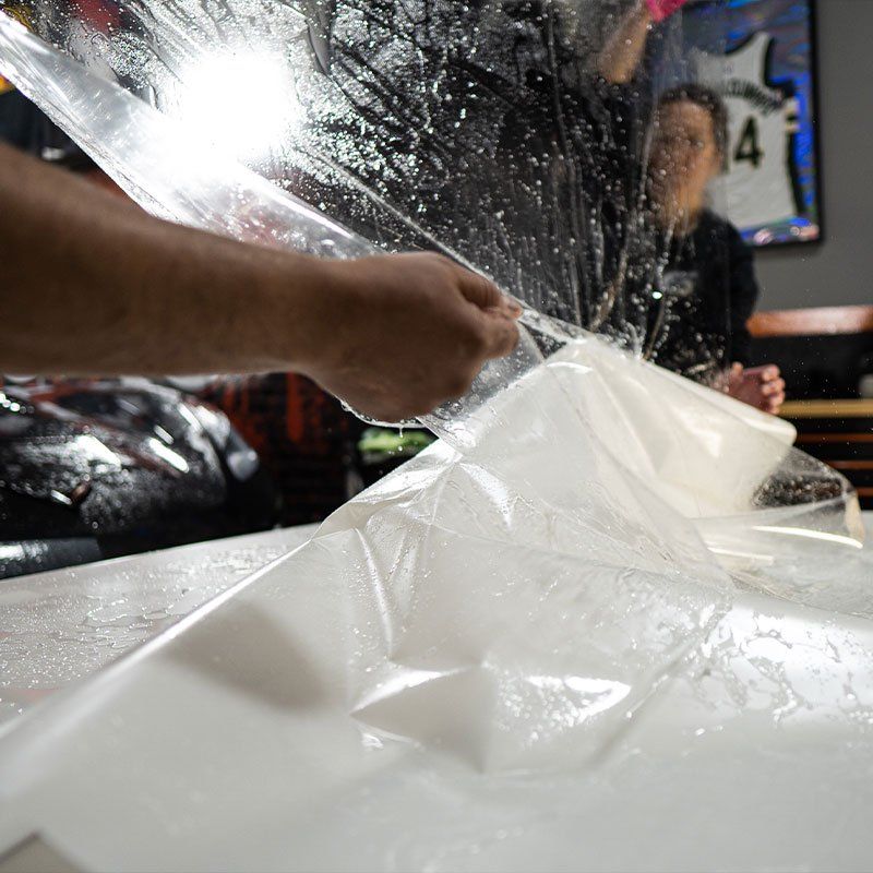 Person peeling protective film from a car surface. Another person is in the background. White, shiny surface.