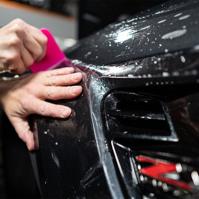 Person using a pink squeegee to apply clear protective film to a black car bumper.