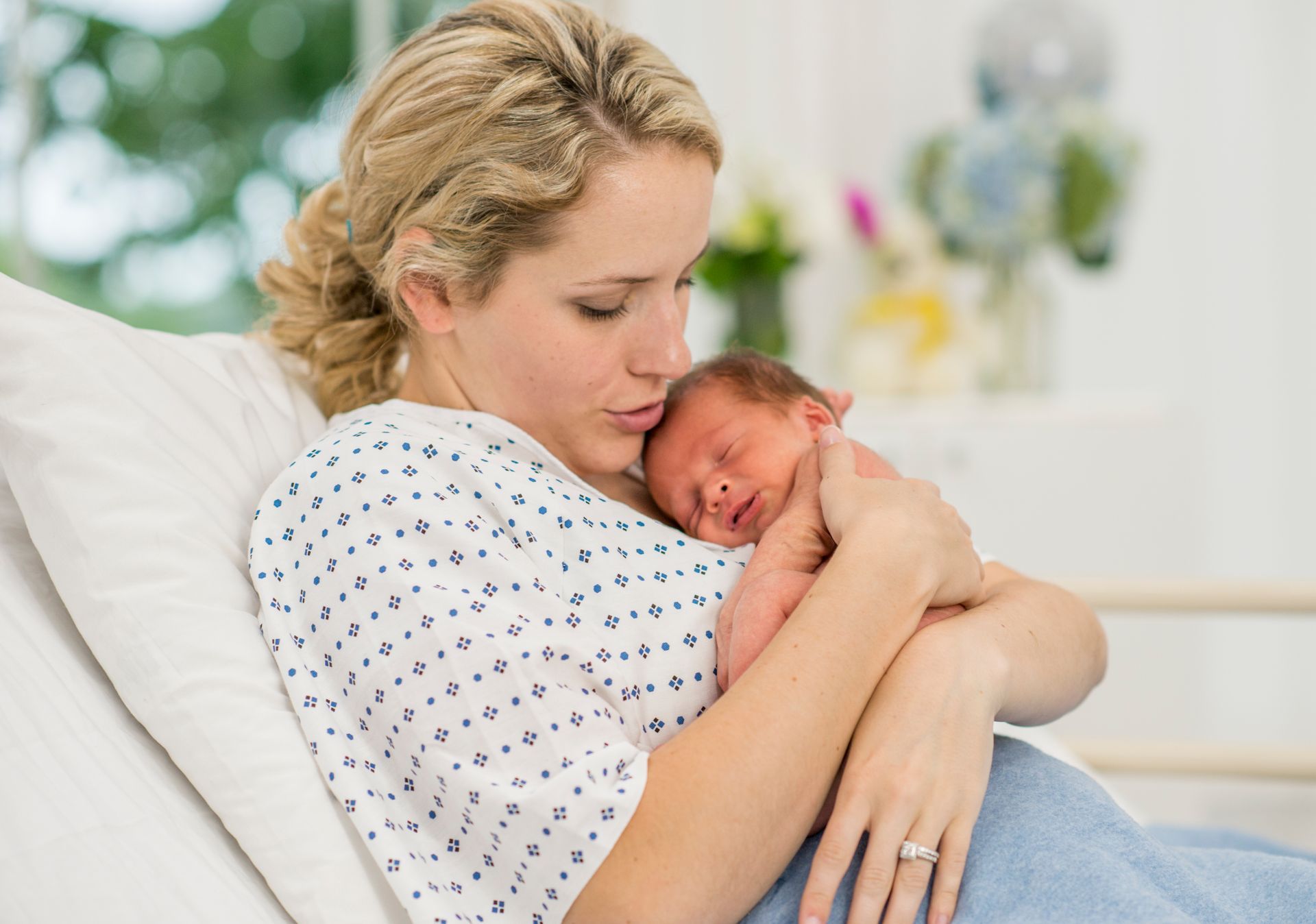 Woman holding and gently cradling a sleeping newborn on a hospital bed, wearing a patterned gown