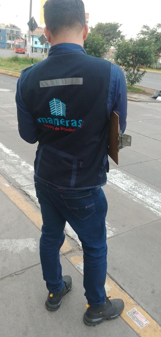 Una persona con uniforme azul marino y un logotipo turquesa camina por el arcén mojado de una carretera, vista desde atrás.