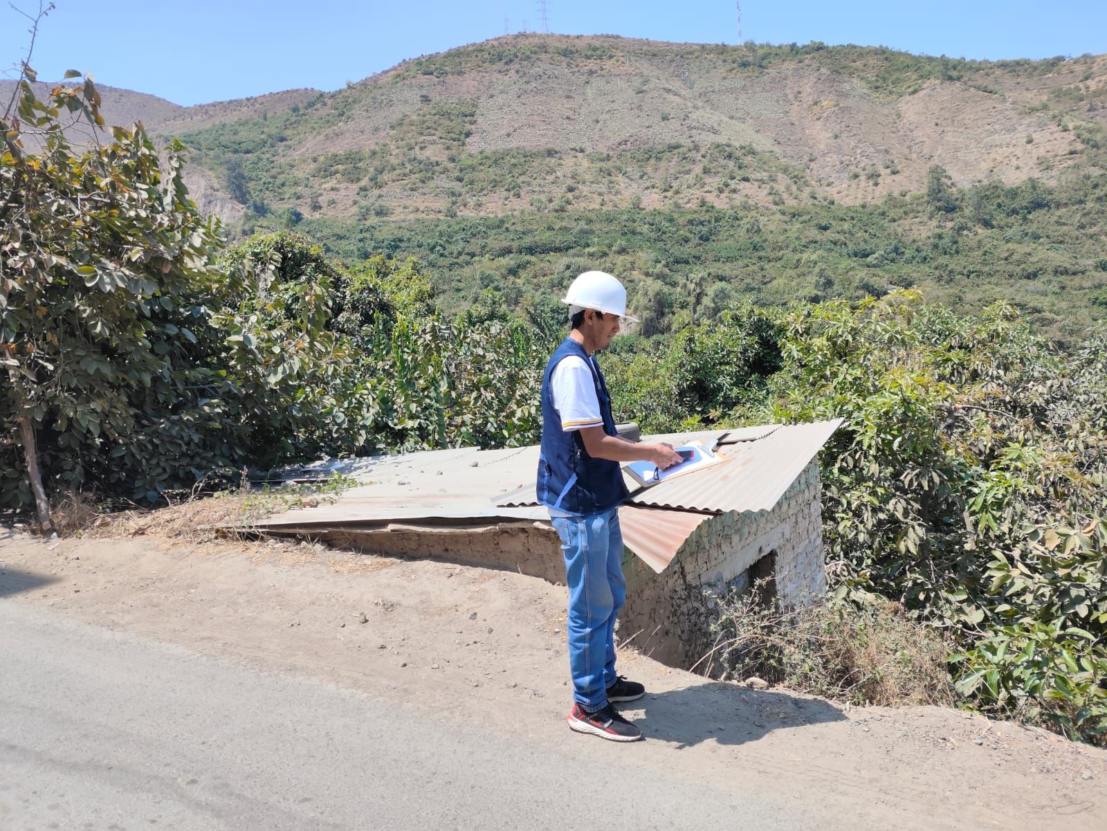 Un trabajador con casco inspecciona una estructura de hormigón al borde de la carretera en una zona montañosa.