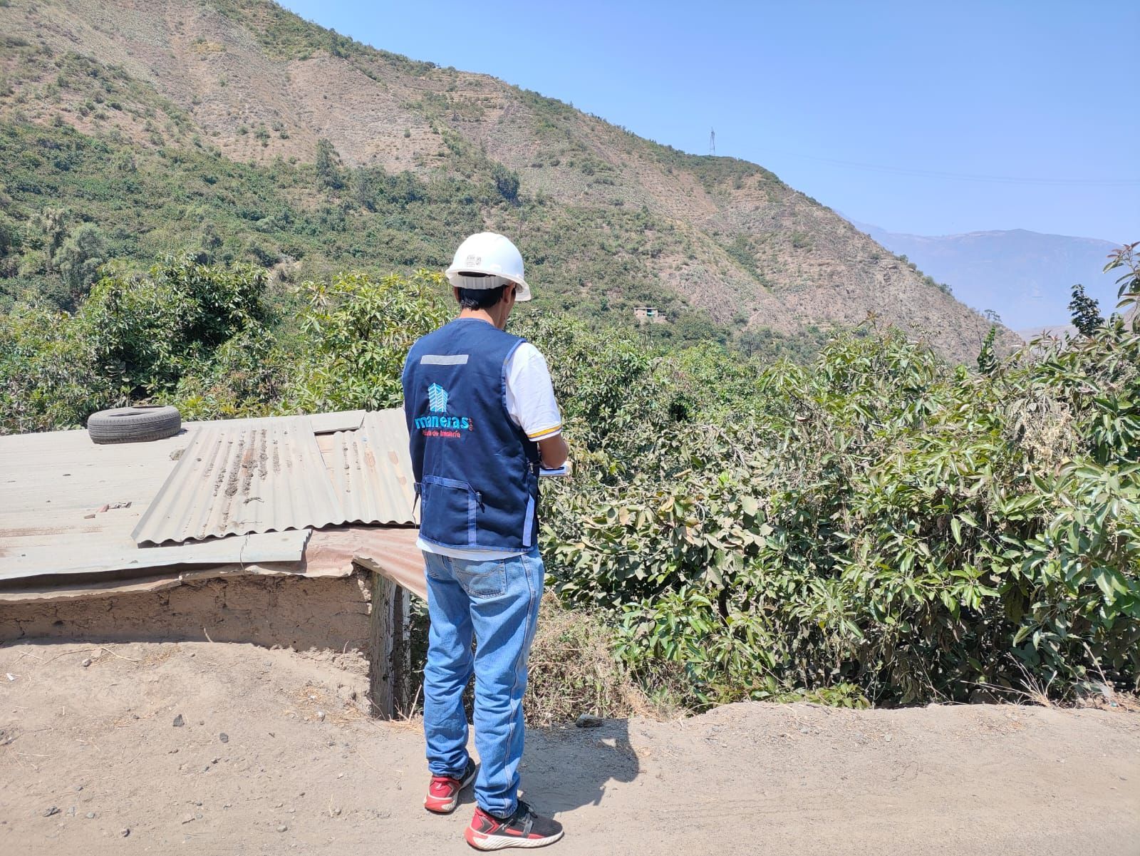 Una persona con casco blanco y chaleco azul se encuentra de pie en un mirador de tierra sobre un valle de montaña verde.