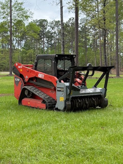 Red skid-steer loader lifting a large uprooted tree stump in a wooded area under a blue sky
